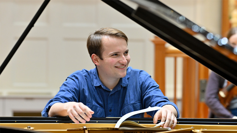 Misha Kaploukhii - A man, wearing a blue shirt, sitting at a piano, smiling to someone off camera.